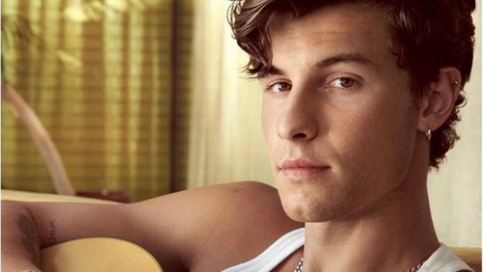 Shawn Mendes posando junto a una guitarra mientras usa una delgada playera en color blanco